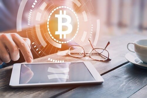 Evertas Acquires Crypto Mining Insurance Pioneer Bitsure Person at a table using a tablet with a graphic of the bitcoin logo hovering above the tablet. Glasses and a cup are also sitting on the table